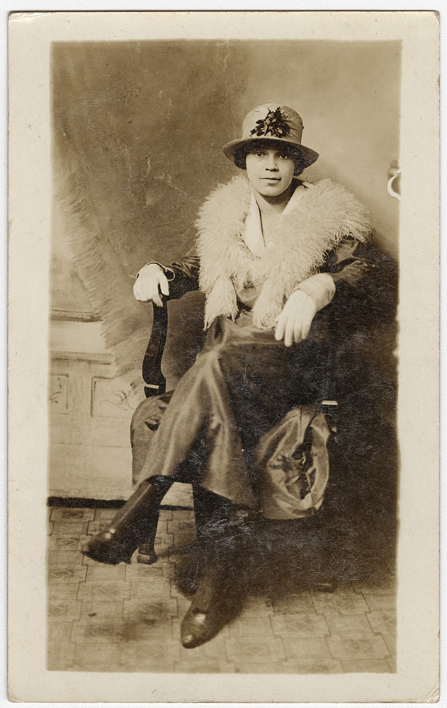 Sepia photograph of a young Black woman seated in a chair, wearing a nice dress, fur stole and cloche hat.