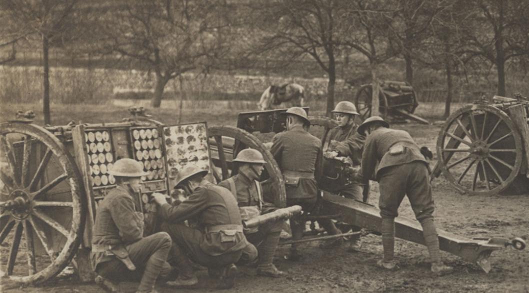 Sepia photograph of six young men wearing WWI uniforms and helmets gathered around a field artillery gun in a grassy area bounded by trees.