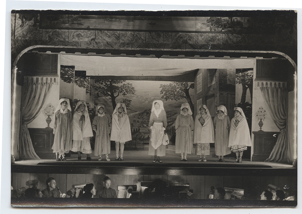 Black and white photograph of nine people wearing nursing uniforms performing on a stage. The orchestra pit in front of the stage is full of musicians. 