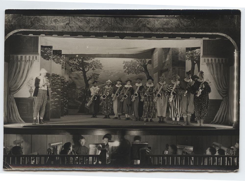 Black and white photograph of ten people dressed as clowns each playing the saxophone on stage. Another person dressed in plaid pants plays the clarinet. 