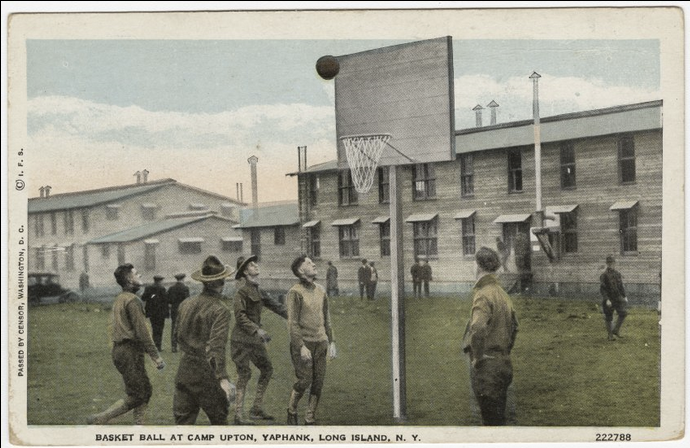 Colorized photograph of young white men in military uniform playing basketball on a grassy field between barracks.