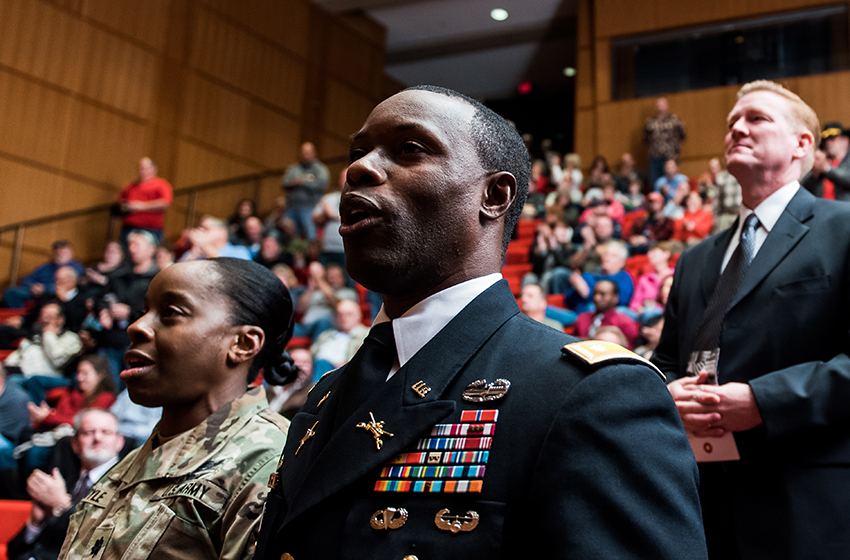 Modern photograph of an auditorium filled with people. In the foreground, a Black man and a Black woman in military uniform are singing something.