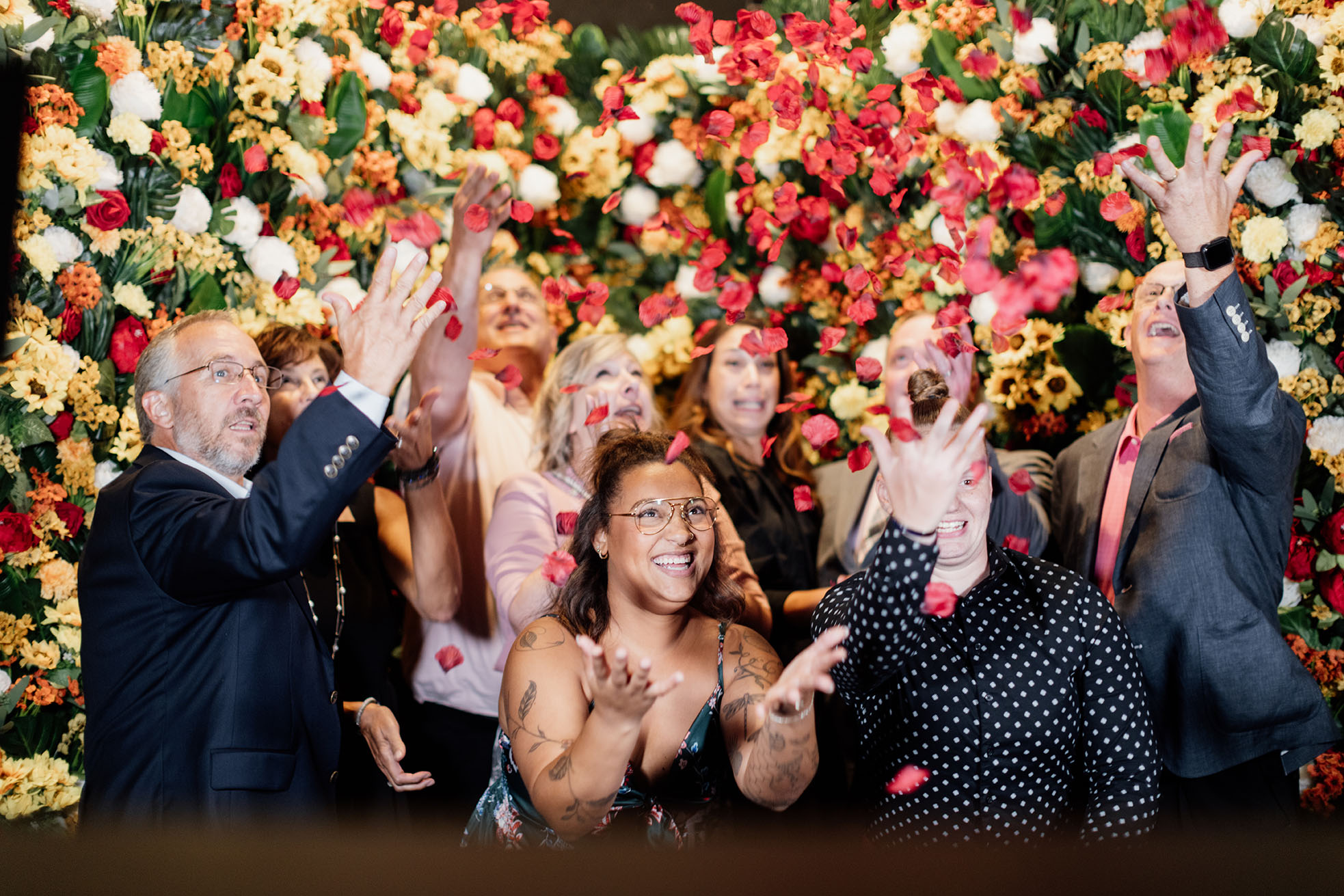 Modern photograph of a group of people standing in front of a flower wall, tossing flower petals up into the air