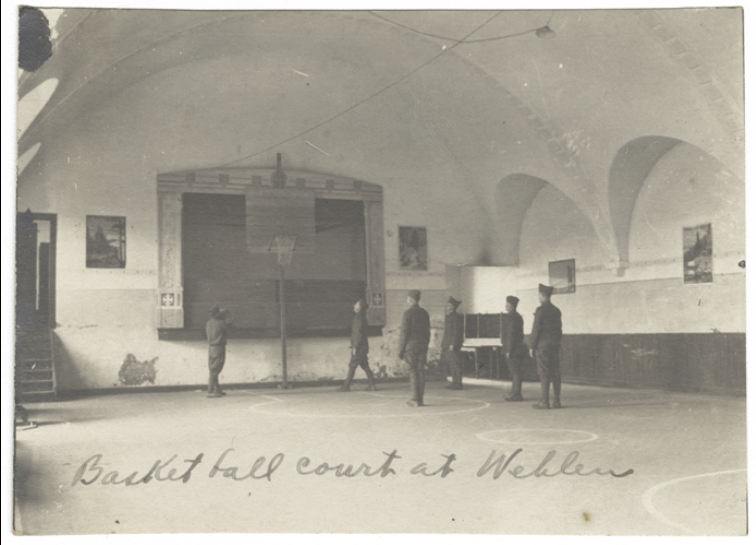 Black and white photograph of a gymnasium hall with a basketball hoop at the far end. Several young men in military uniform are playing basketball.