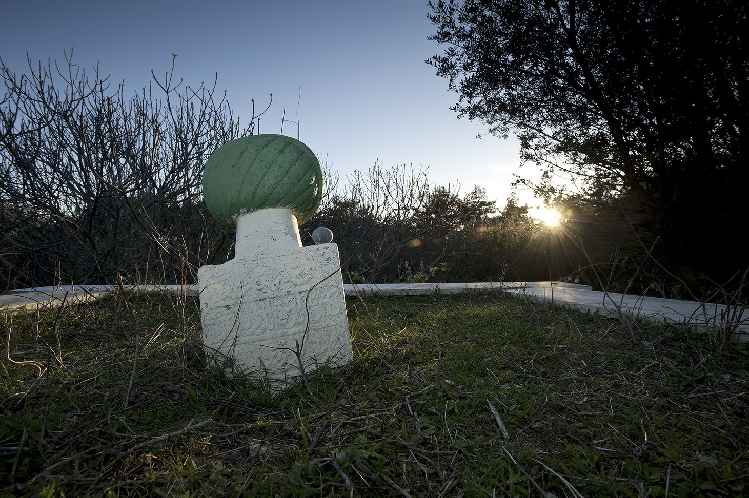 Modern photograph of a lone grave marker topped by a turquoise stone in the foreground as the sun sets in the background
