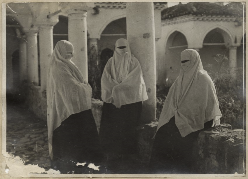 Black and white photograph of three people standing and seated in an open-air courtyard bounded by arches and pillars. The people are wearing dark skirts and light-colored tops and head coverings.