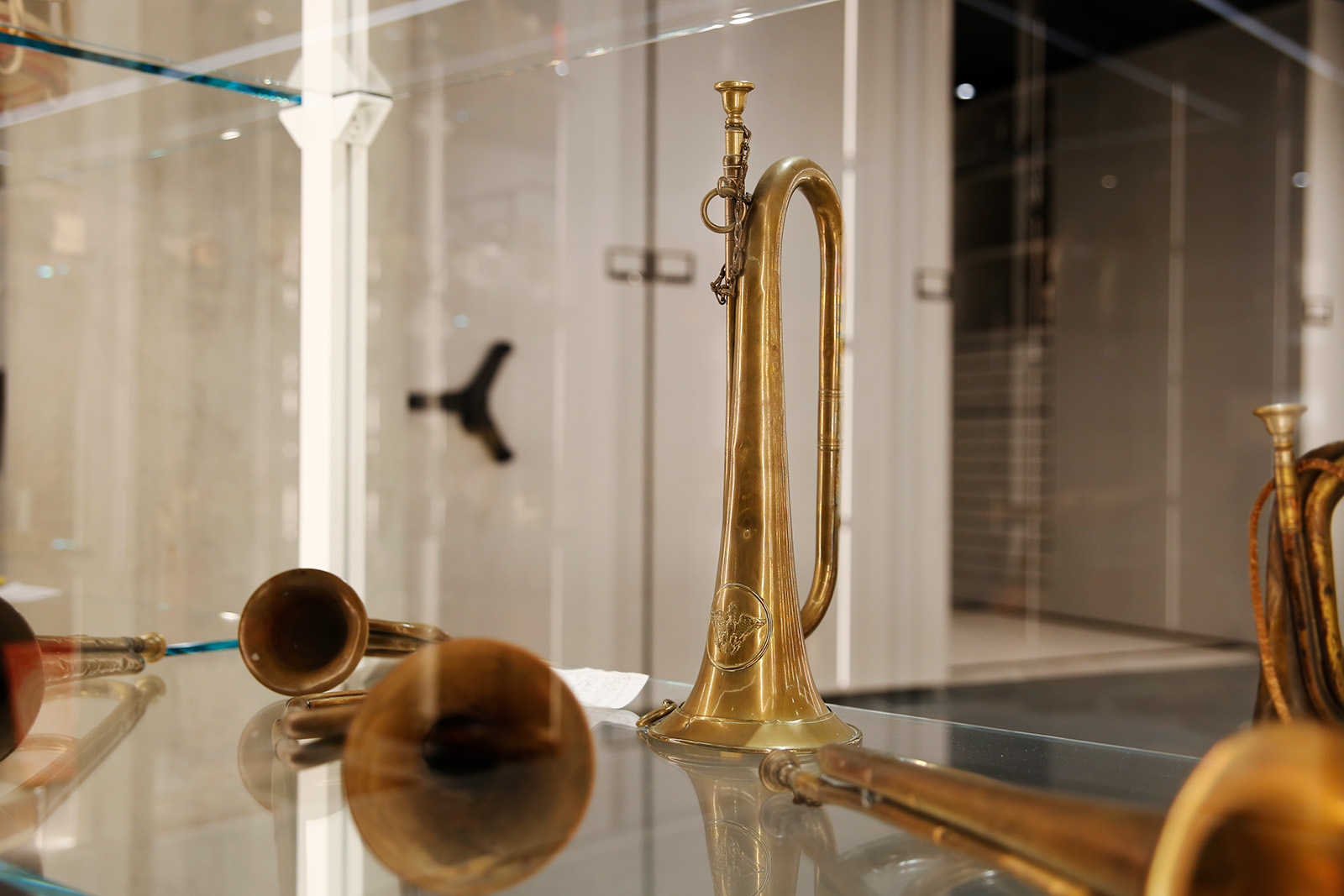 Modern photograph of a glass display case featuring a bugle resting upright on a glass shelf surrounded by more bugles