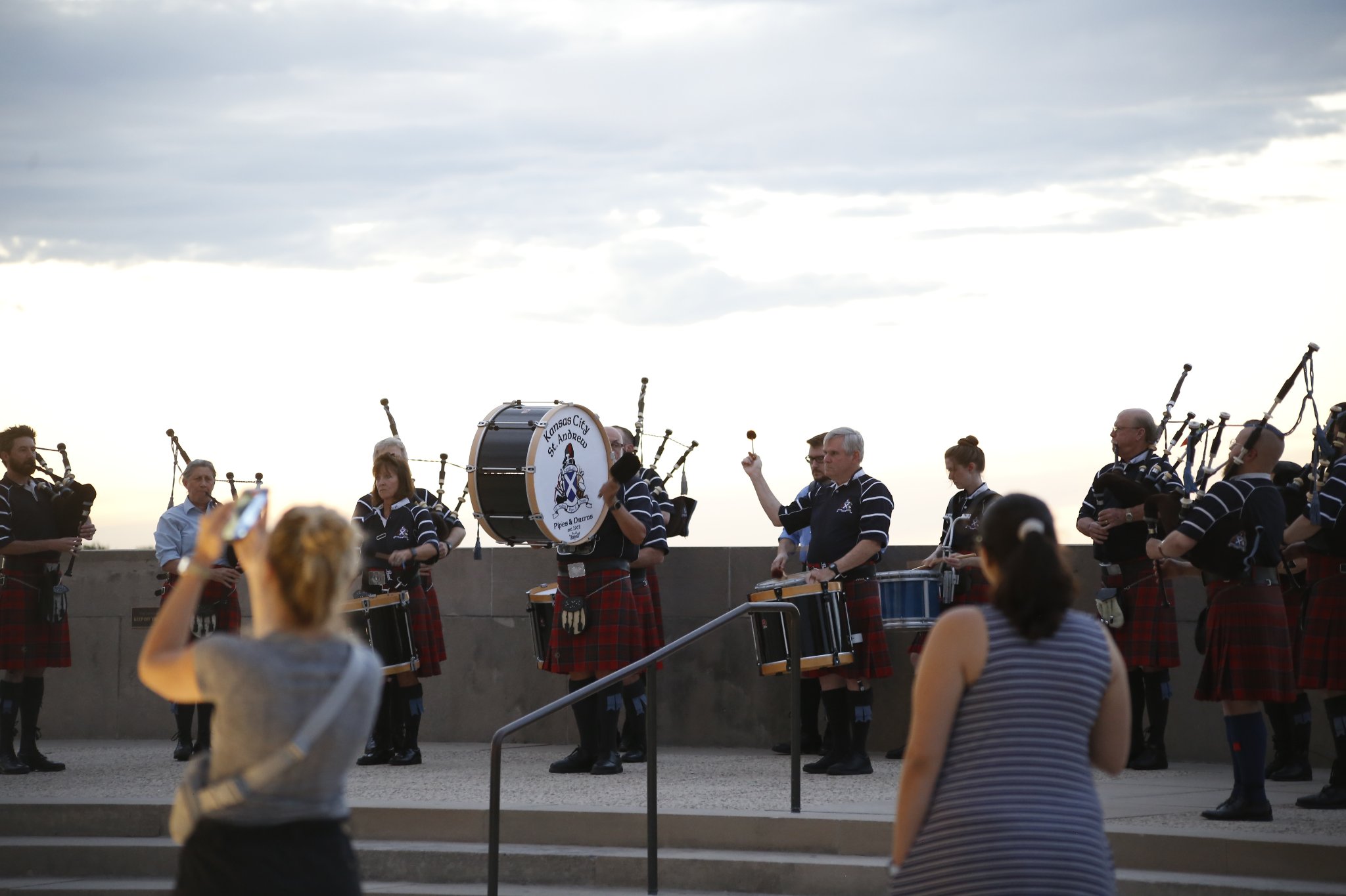 Modern photograph of a large band of people wearing kilts standing on Memorial Courtyard. They are playing a variety of drums and bagpipes. Onlookers take photos on them.