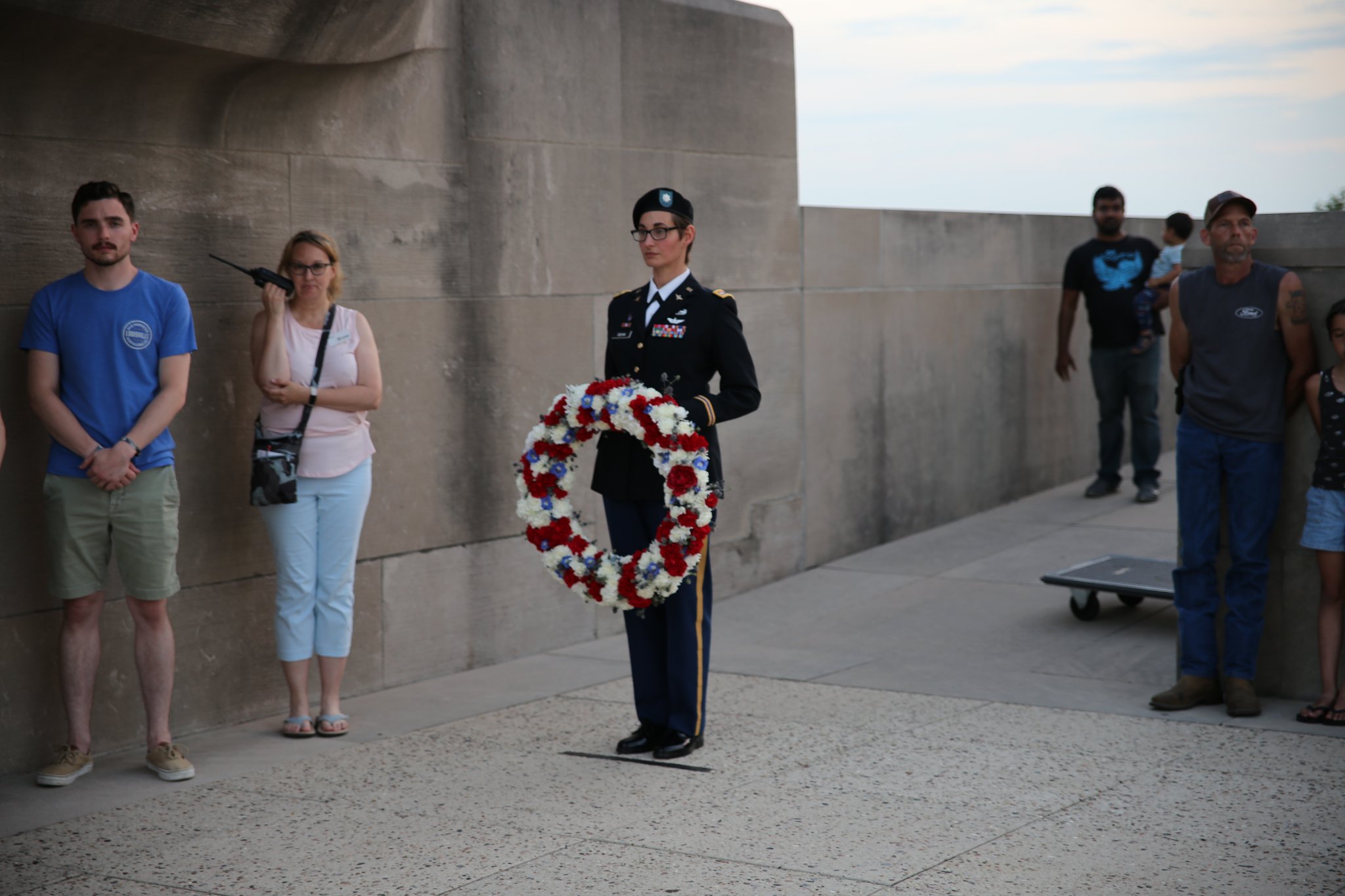 Modern photograph of a stone courtyard. Several people in civilian clothes are gathered around a person in military uniform. The military person is holding a large floral wreath.