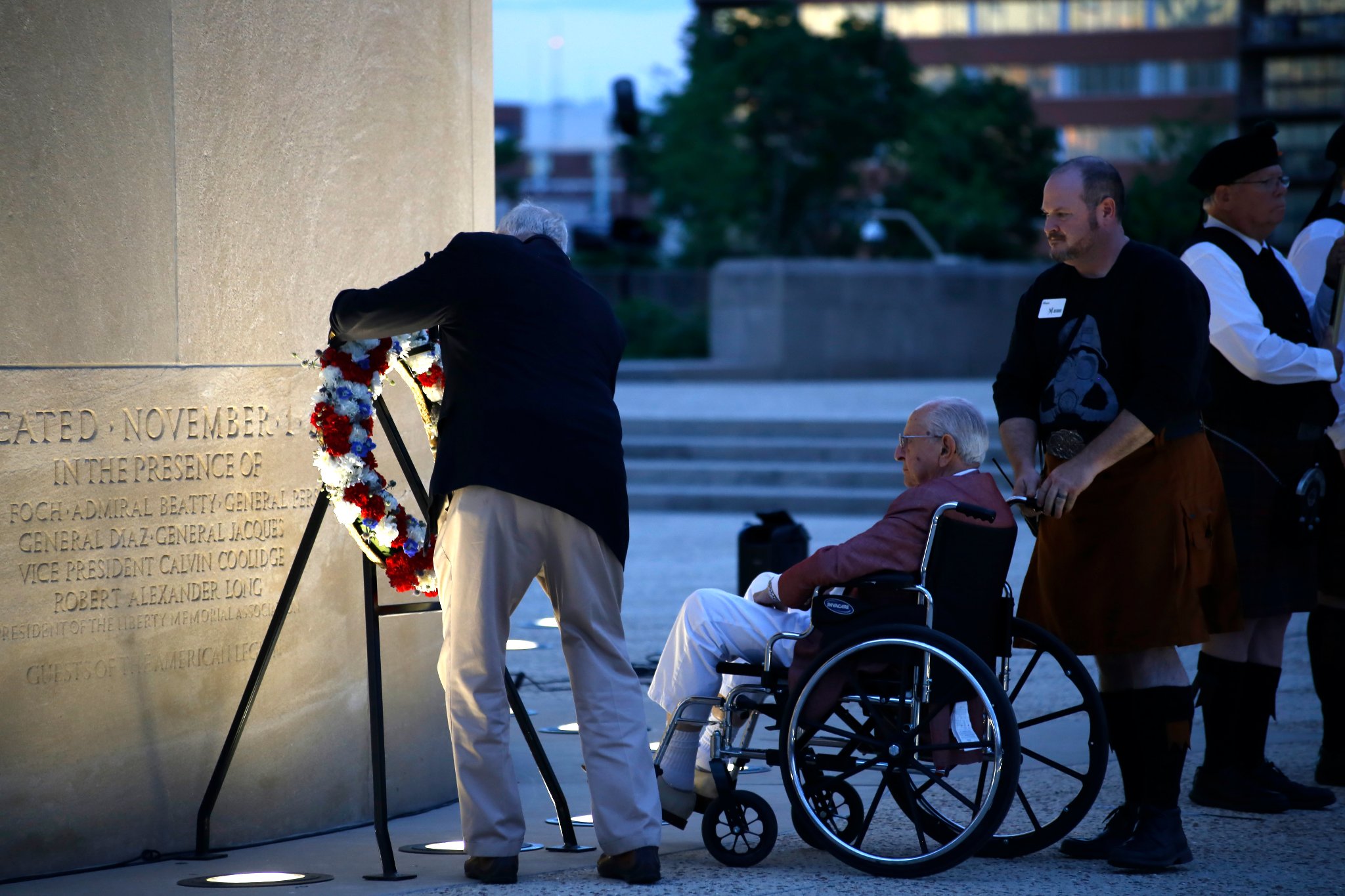 Modern photograph of the base of the Liberty Memorial Tower at twilight. A person in a dark jacket places a large floral wreath on a stand. Next to them, an older person observes while seated in a wheelchair being pushed by a man wearing a kilt.