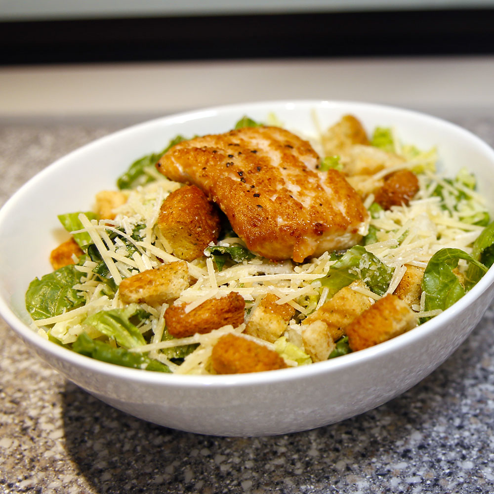 Modern photograph of a lettuce salad with croutons and shaved parmesan cheese topped with a breaded and fried hunk of chicken. 