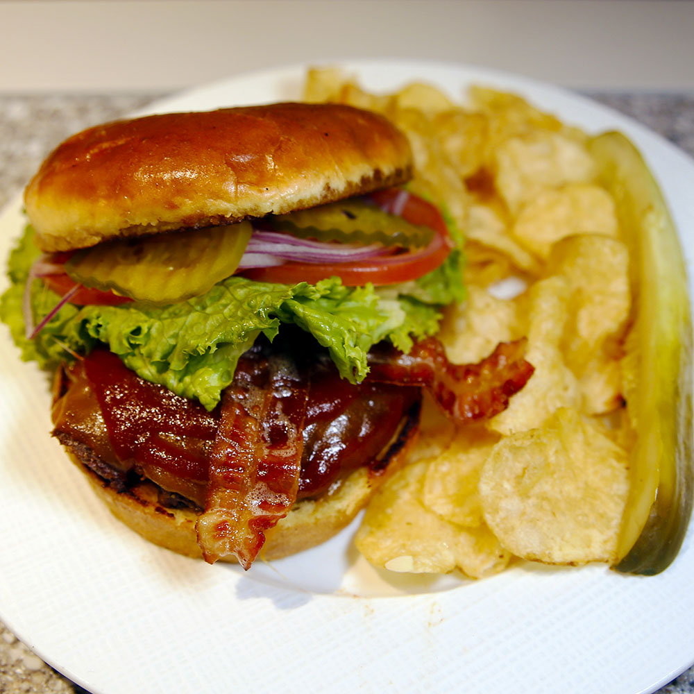 Modern photograph of a burger with bacon, lettuce, tomatoes and sliced pickles, accompanied by potato chips and a long pickle slice.