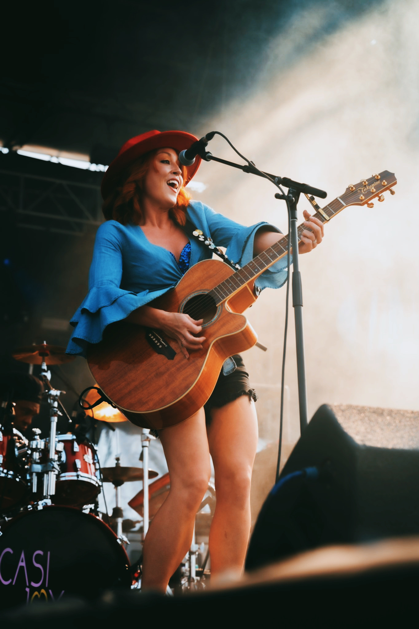 Modern photograph of a red-haired white woman wearing a red wide-brimmed hat and a blue shirt with bell sleeves. She is playing the guitar and singing into a microphone.
