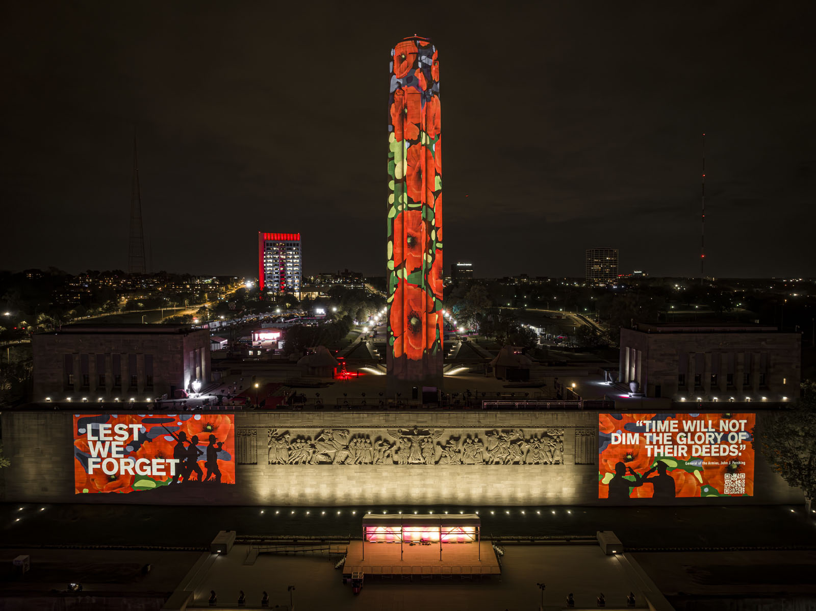 Modern photograph of the Liberty Memorial Tower, Great Frieze and North Terrace Wall taken from high above the North Lawn. The Tower is covered in projections of poppies. More poppies plus text and pictures are projected onto the North Terrace Wall on either side of the Great Frieze.
