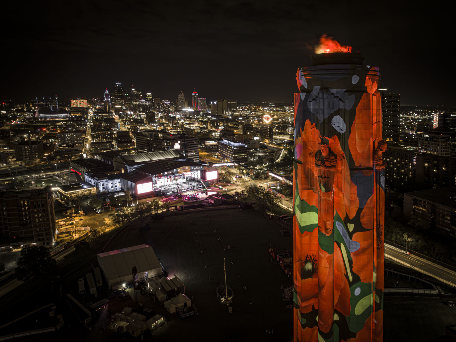 Modern photograph taken high in the air about level with the top of the Liberty Memorial Tower. The Tower is in the foreground with the carved stone guardians visible, covered in poppy projections. In the background, the NFL Draft Stage in front of Union Station is lit up.