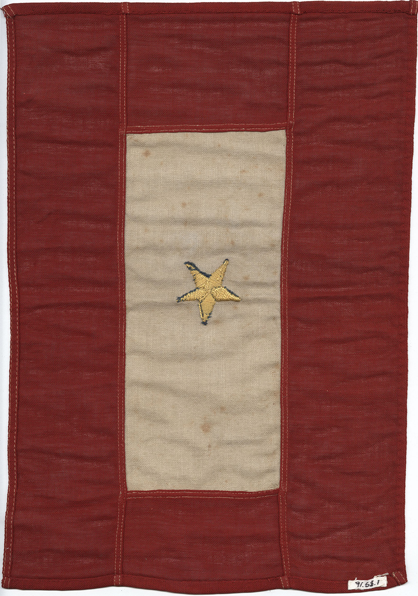 Photographie moderne d'un drapeau de la Première Guerre mondiale. Une bordure rouge entoure un rectangle crème/blanc. Une seule étoile dorée est brodée au centre, avec des bords bleus montrant qu'une étoile bleue est en dessous.