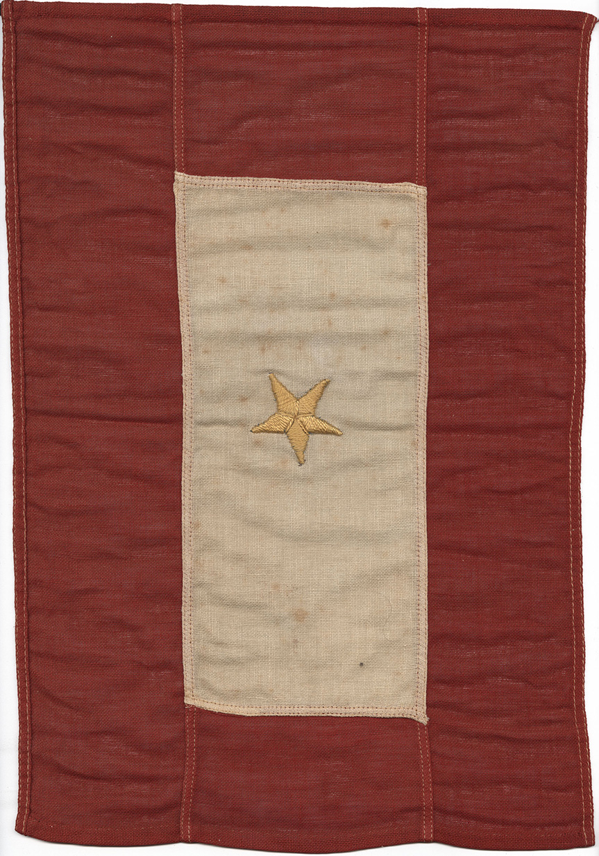 Photographie moderne d'un drapeau de la Première Guerre mondiale. Une bordure rouge entoure un rectangle crème/blanc. Une seule étoile dorée est brodée au centre.