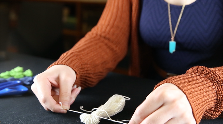 A person tying a second piece of yarn closer to the top of the bundle of looped yarn (side view)