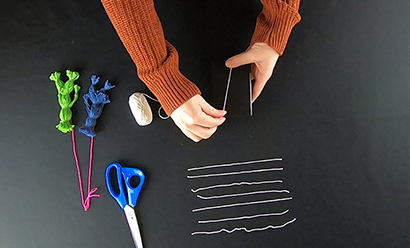 Top down shot of a table with yarn, scissors and completed yarn dolls. A person (only the arms are visible) is wrapping yarn around a small book.