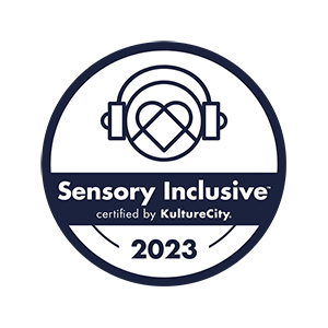 White circular logo with a dark blue outline. Inside the circle is a simple graphic of headphones being "worn" by a smaller circle with a graphic heart inside. Text inside logo: "Sensory Inclusive / certified by KultureCity / 2023"