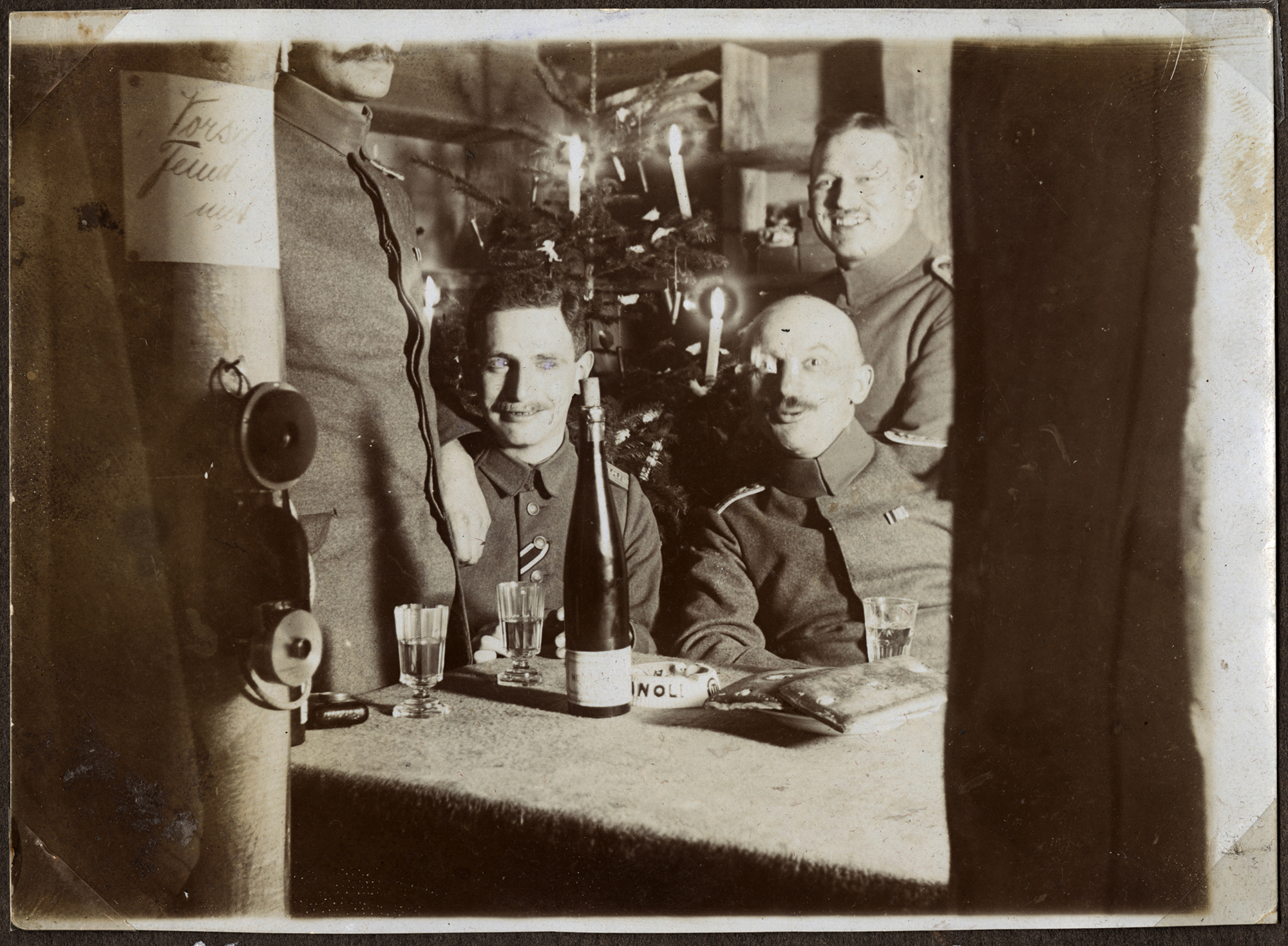 Black and white photograph of several WWI soldiers sitting in front of a Christmas tree with smiles on their faces and drinks on the table