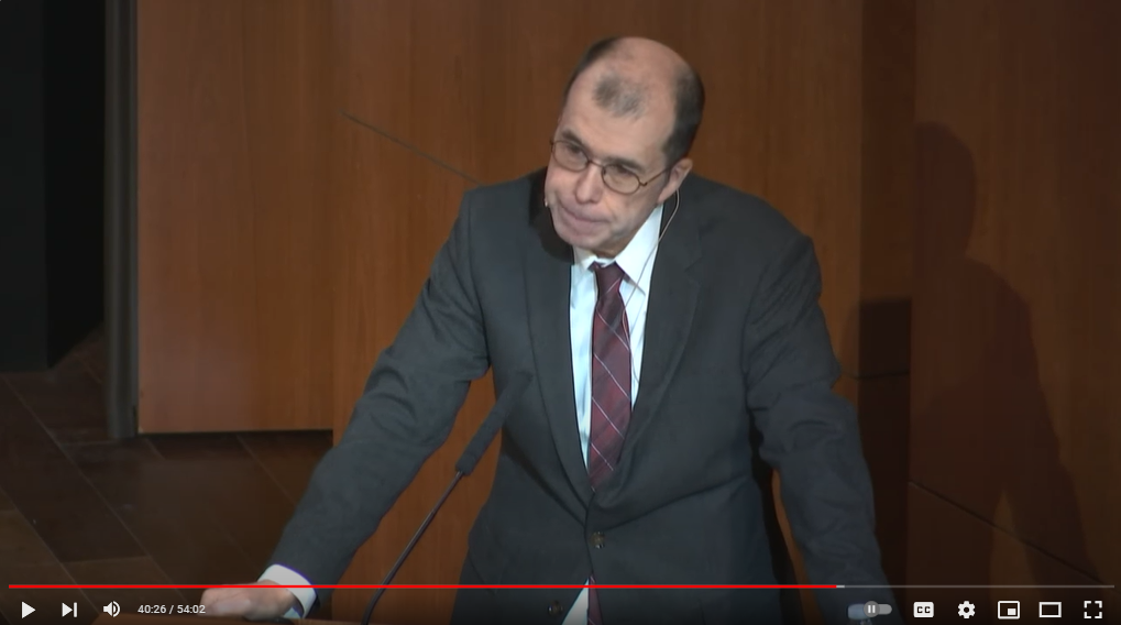Capture d'écran d'une vidéo de conférence. Un homme blanc en costume gris parle sur un podium.