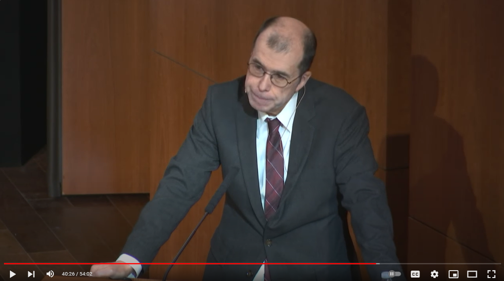 Screenshot of a lecture video. A white man in a gray suit speaks while at a podium.