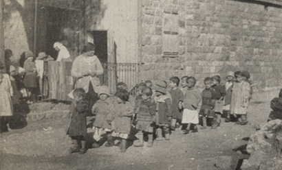 Black and white photograph of children walking on a street. A woman stands with the group.