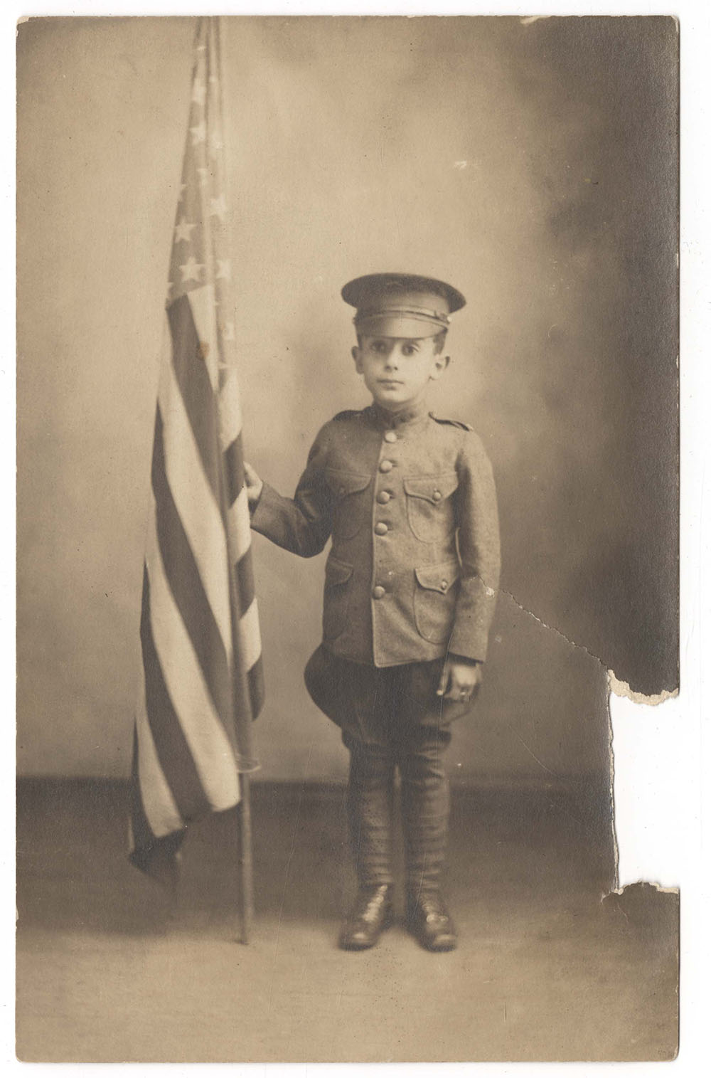Sepia photograph of a small white boy dressed in a play military uniform and cap, holding a United States flag that is taller than he is.