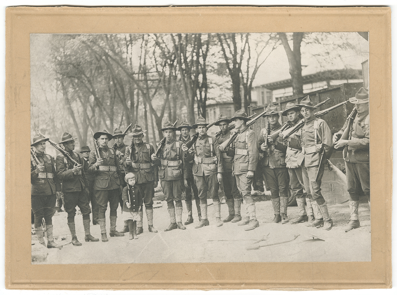 Black and white photograph of a group of soldiers standing in a line with rifles on their soldiers in an outside field. A little white girl in a dress and coat stands in front of them with a toy rifle on her shoulder. The soldiers are laughing and smiling indulgently at her.