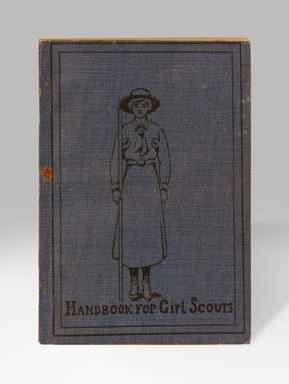 Closed book with a dark grey cover illustrated with a line drawing of a girl wearing a uniform hat, shirt and skirt. Text: 'Handbook for Girl Scouts'