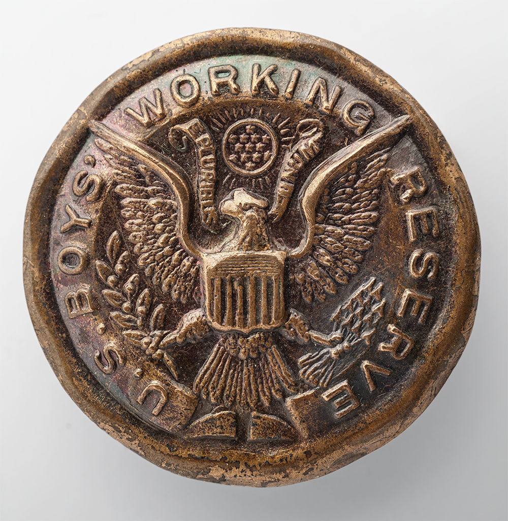 Modern photograph of a circular bronze-colored pin embossed with a bald eagle holding olive branch and arrows. Text running around the edge: 'U.S. Boys' Working Reserve'