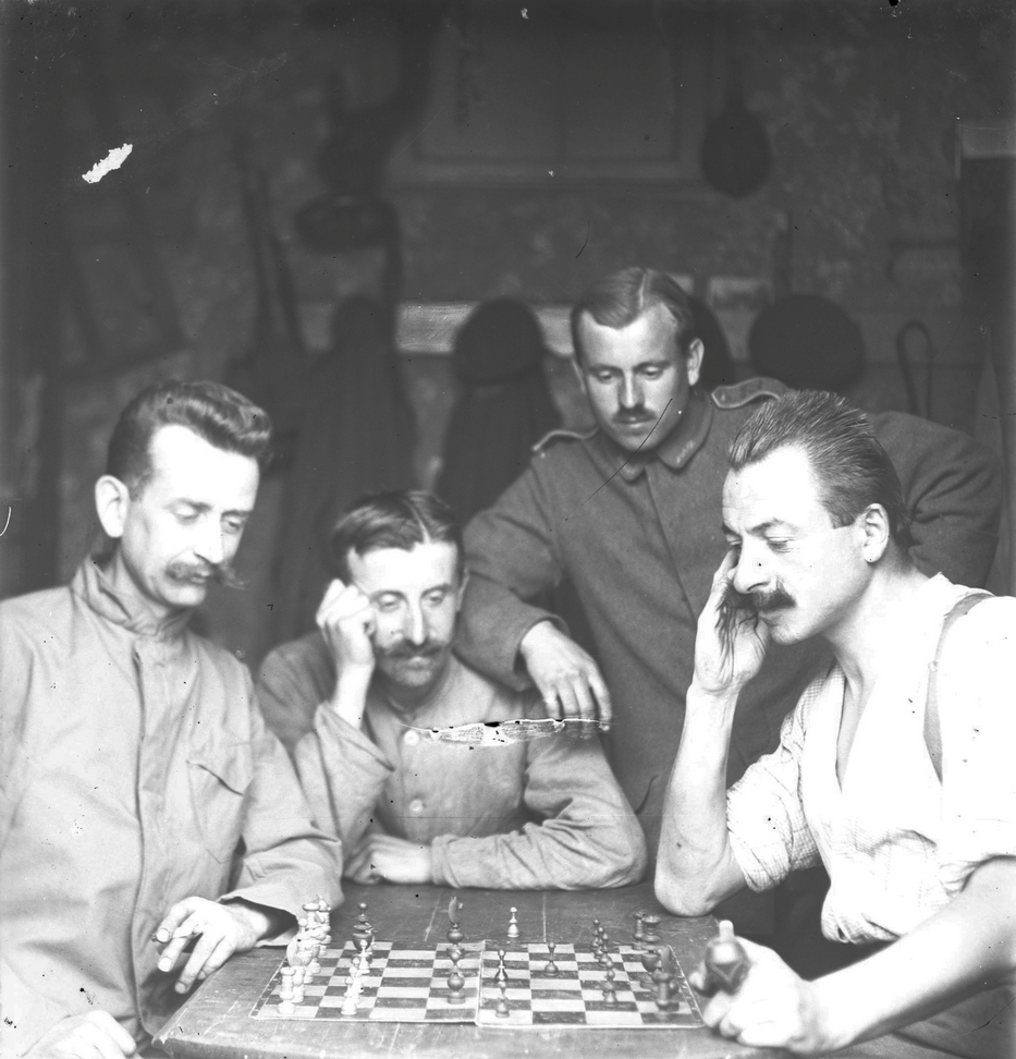 Two German soldiers play chess while two others look on. Coats are hung up on hooks behind them. All four have full, pointy mustaches.