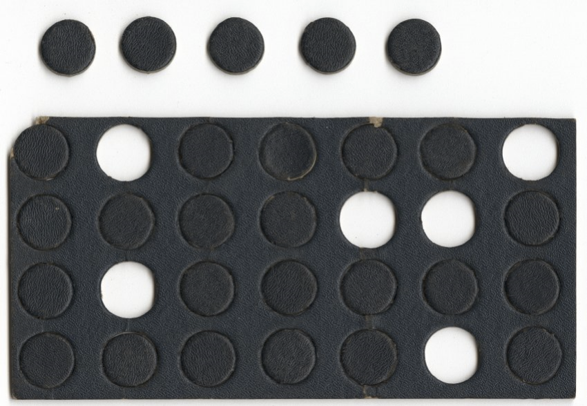 Modern photograph of a rectangular piece of thick cardboard coated in black paint, perforated with many circles. Some of the circles have been snapped out and sit to the side. 