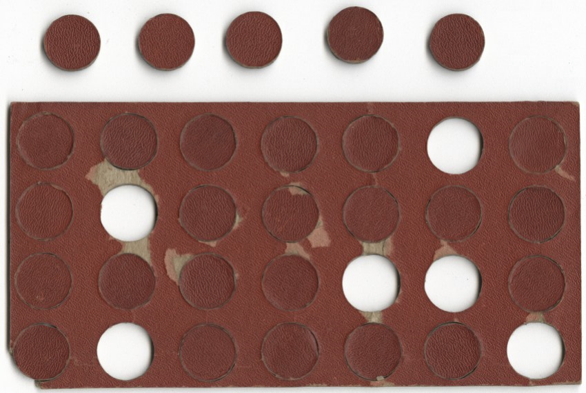 Modern photograph of a rectangular piece of thick cardboard coated in red paint, perforated with many circles. Some of the circles have been snapped out and sit to the side.