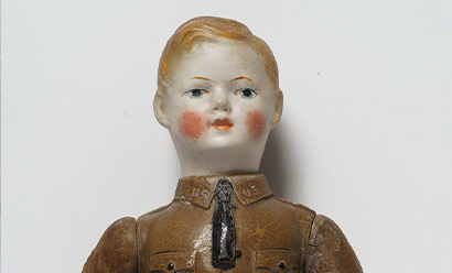 Modern photograph of a WWI-era doll shaped like a white boy with short blond hair wearing a military uniform shirt and tie.