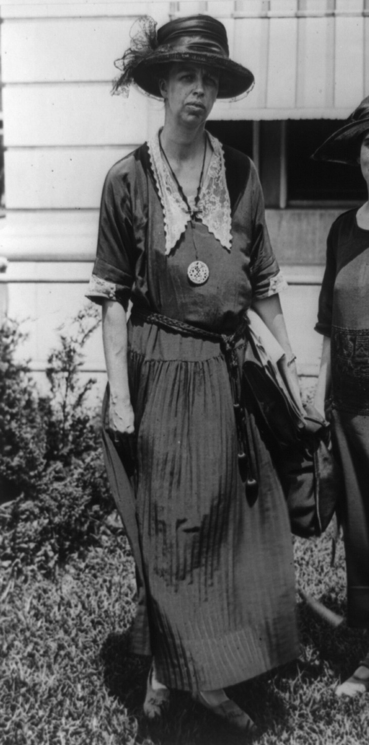 Black and white photo of a tall white middle-aged woman standing on a lawn outside a white clapboard building wearing a simple walking dress and feathered hat.