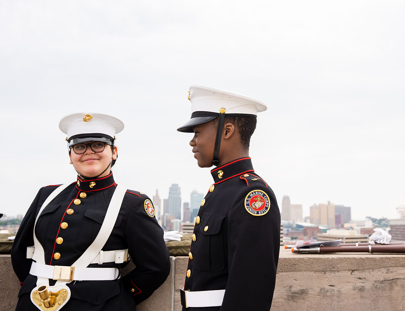 Modern photograph of a white female teenager and a Black male teenager in Junior ROTC dress uniforms and hats, in a candid smiling moment on Memorial Courtyard.