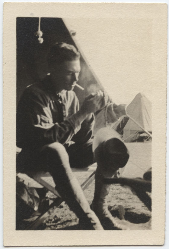 Black and white photo of a young white man in U.S. military uniform seated outside a tent, lighting the cigarette in his mouth.