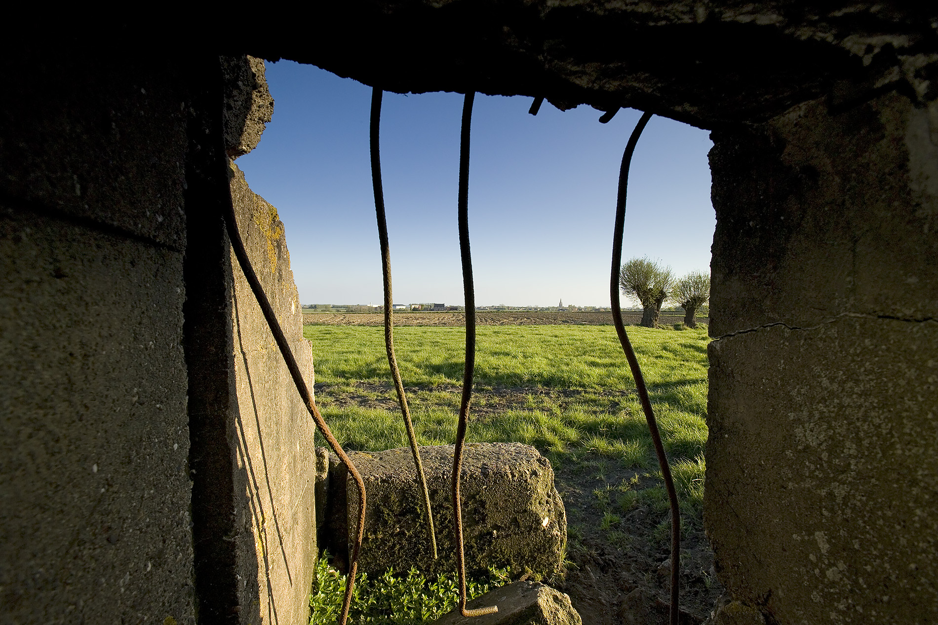 Modern photograph from the inside of the remains of a concrete bunker, looking through twisted iron struts out to a grassy field on a sunny day.