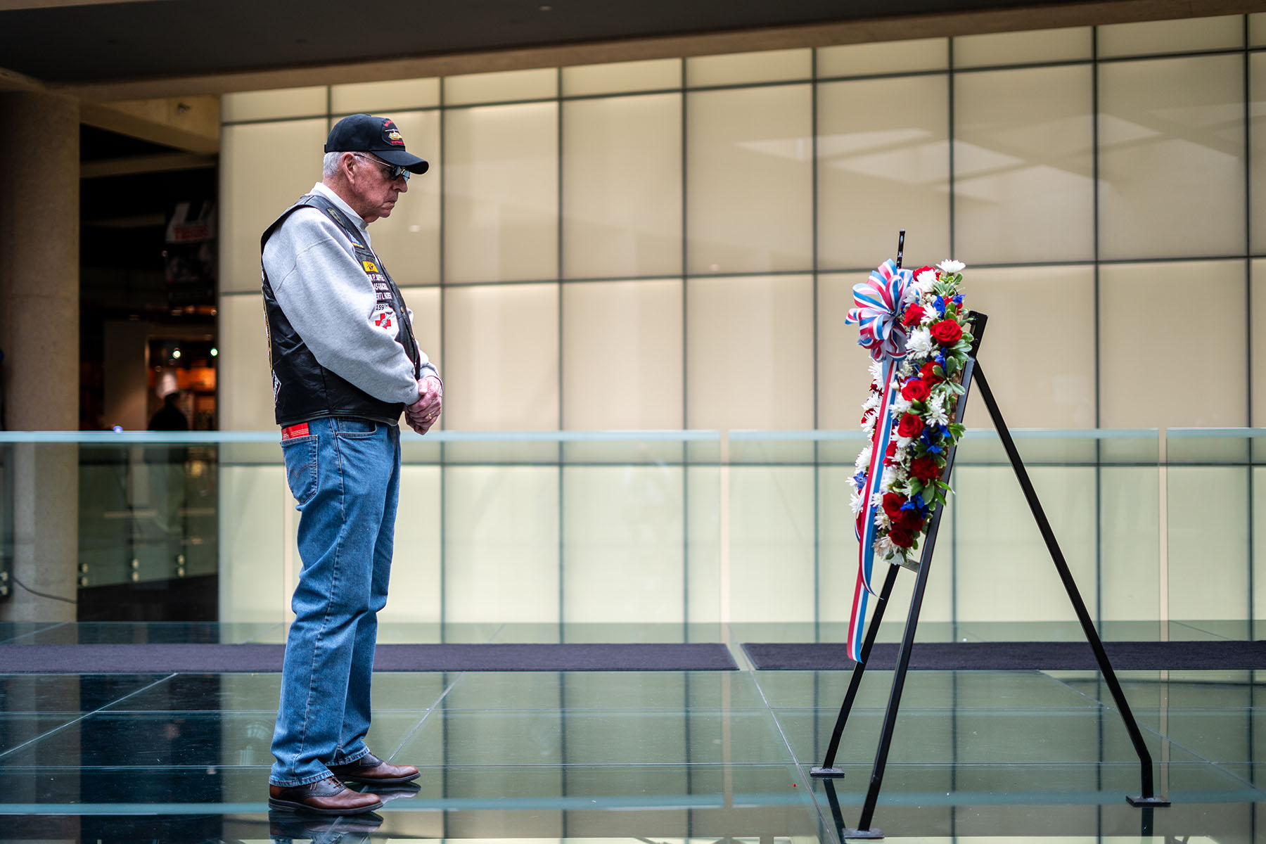 Modern photograph on the Glass Bridge in the Museum. An older white man wearing a veterans' baseball hat and vest stands and contemplates a remembrance wreath displayed on the bridge.