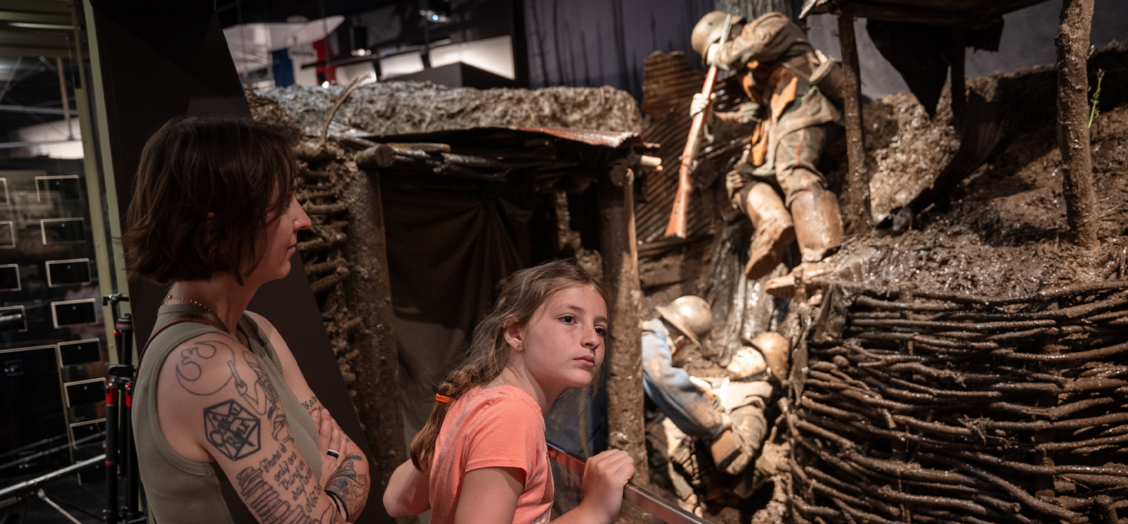 Modern photograph of a museum exhibit recreation of a trench, with soldier mannequins fighting hand-to-hand in it. Two museum guests, a white adult woman and a white girl dressed casually, gaze at the exhibit. The girl is leaning forward to get a better look.