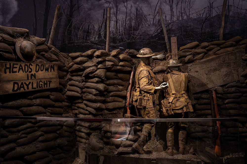 Modern photograph of a museum exhibit recreating a British WWI trench. A soldier mannequin peers through a trench periscope while another soldier mannequin readies to throw a grenade.