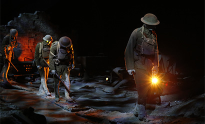Modern photograph of life-size WWI soldier mannequins trudging across a muddy battlescape, lit starkly by orange and white spotlights