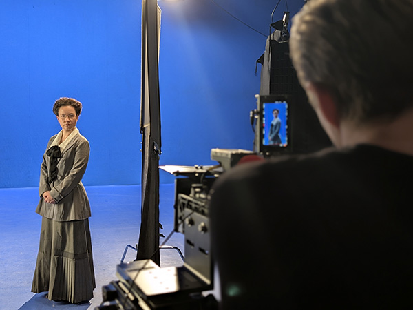 Modern photo of a white female actor in a grey WWI-era skirt and jacket standing in a bluescreened studio being filmed by large elaborate cameras