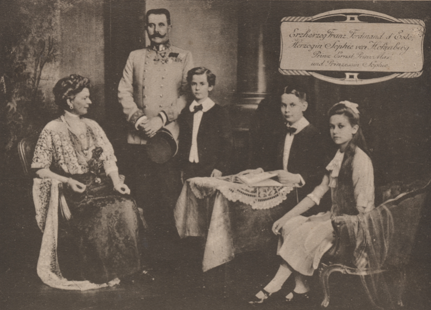 Black and white photograph of Archduke Franz Ferdinand standing beside one of his sons. His wife, Sophie, another of his sons, and his daughter, all sit on couches in the frame. There is a table with a tablecloth in the center.