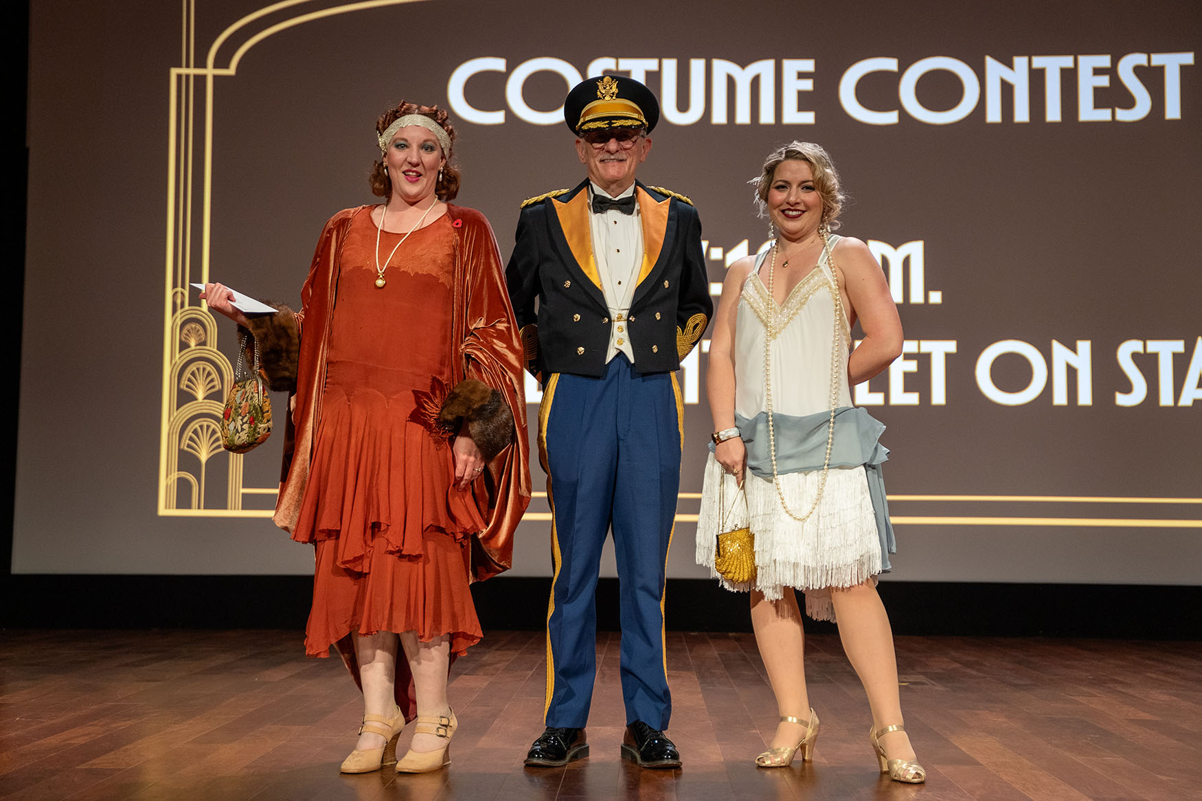 Modern photo of a woman dressed in red-orange flapper attire, a man dressed in vintage military uniform, and a woman dressed in ivory and blue flapper attire posing onstage in the Auditorium