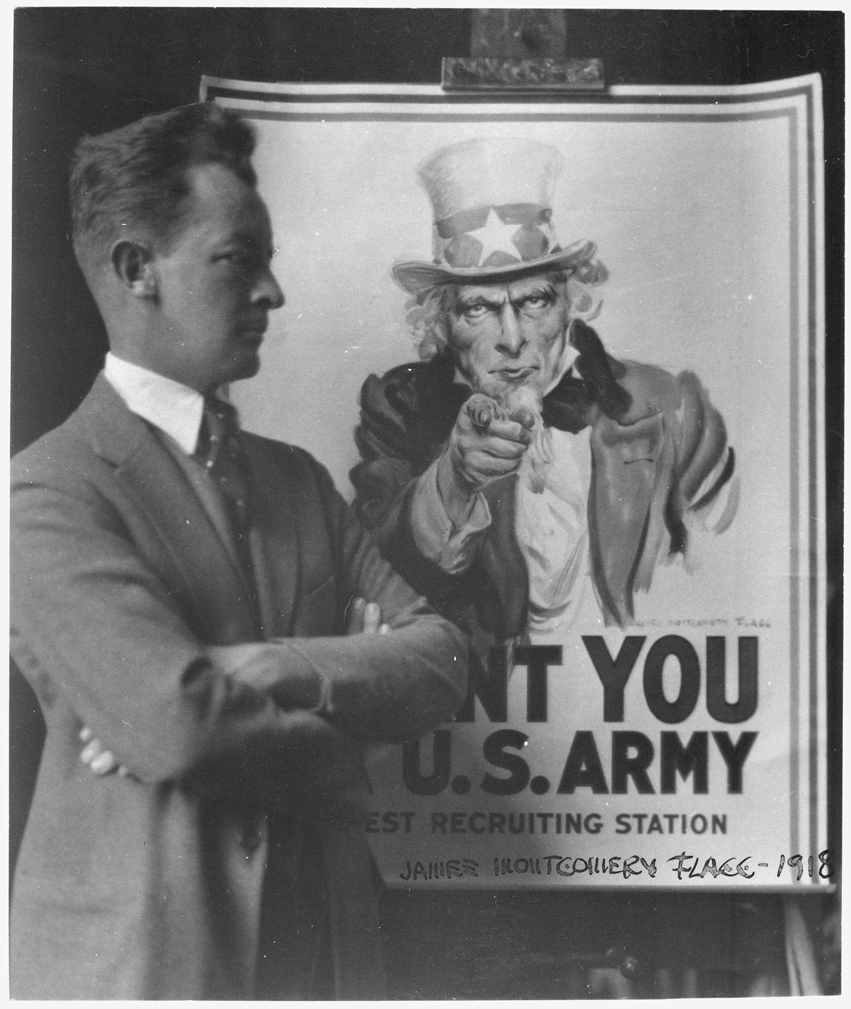 Black and white photo of a thin white man in a suit standing next to an easel displaying the poster of Uncle Sam pointing at the viewer