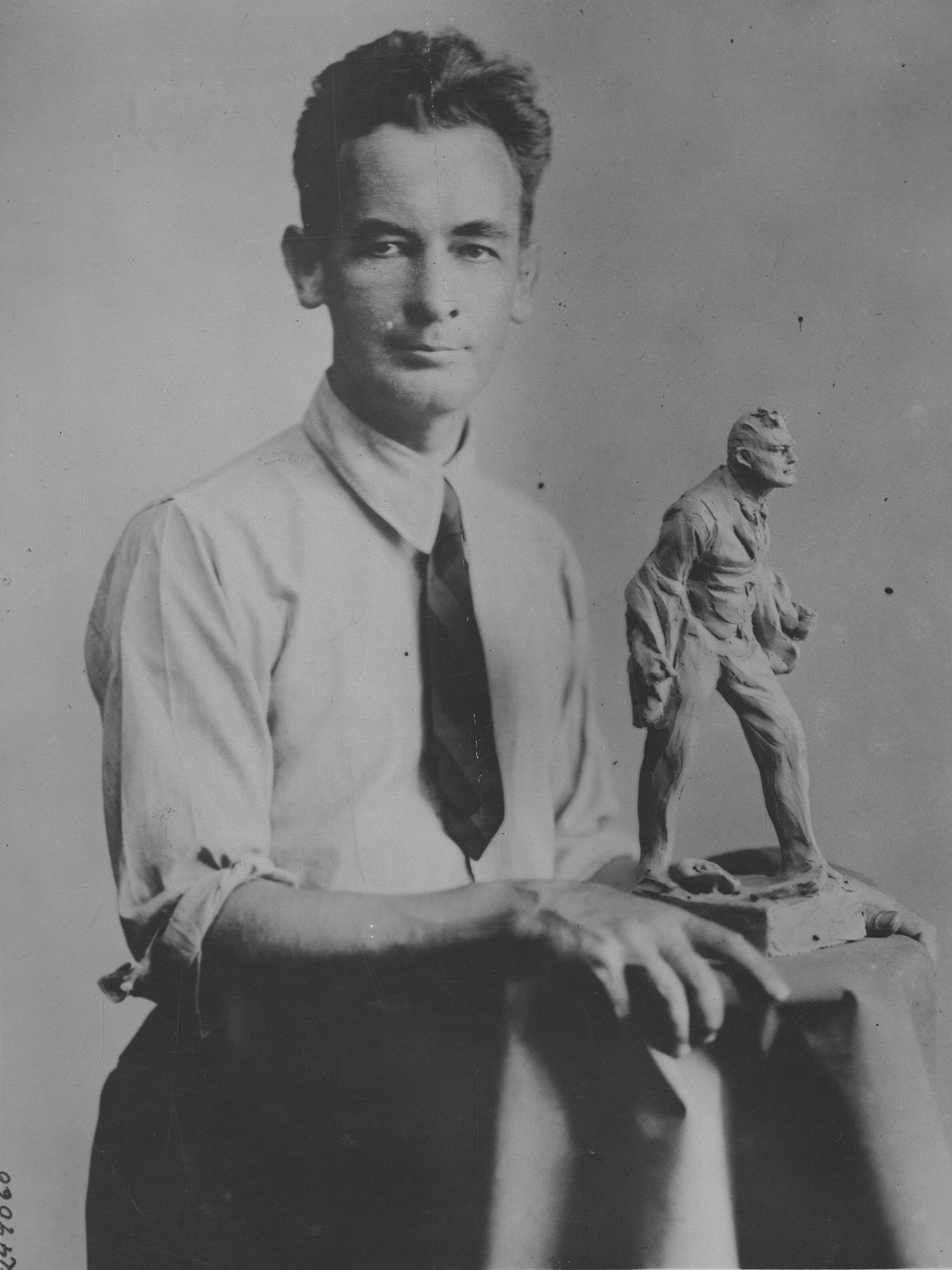 Black and white photo of a thin white man in shirtsleeves and tie posing next to a small statuette of a man taking off his suit jacket