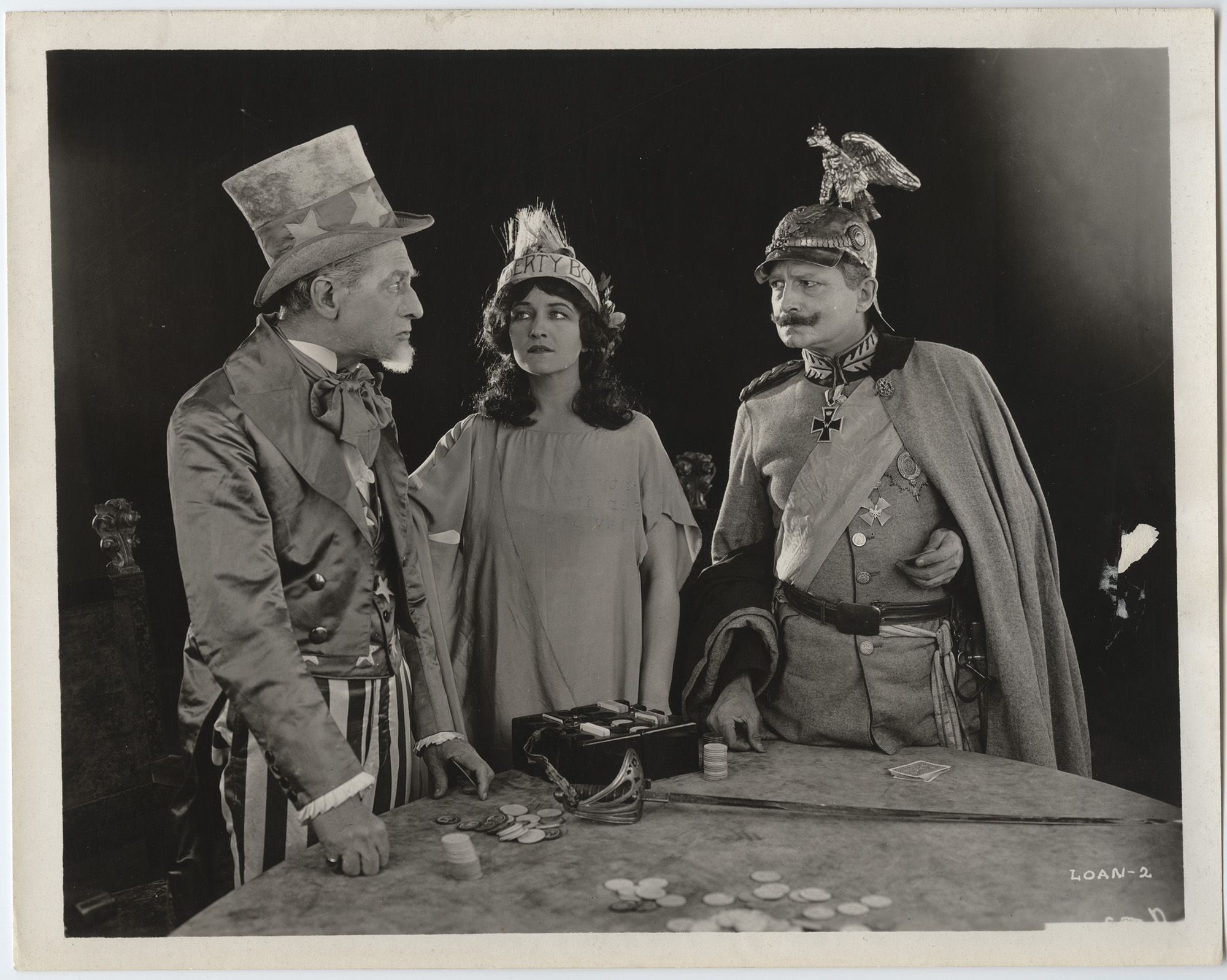 Three actors dressed as Uncle Sam, Lady Liberty, and Kaiser Wilhelm stand around a poker table scattered with poker chips, glaring at each other.